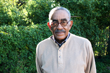 A portrait of a senior, medium-dark skinned man with a mustache wearing round brown glasses and a beige shirt. The man is softly smiling and standing outside in front of greenery. 
