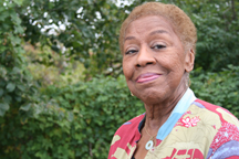 A portrait of a senior medium-dark skinned woman wearing a bright patterned shirt. The woman is smiling and standing outside in front of greenery. 