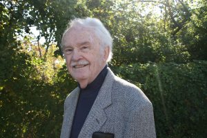 A portrait of a light skinned man with short white hair and a mustache wearing a black turtleneck and suit jacket. The man is smiling and standing outside in front of greenery.