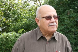 A portrait of a senior medium-dark skinned man with a mustache wearing tinted glasses and a brown collared shirt. The man is looking into the distance and is standing outside in front of greenery.