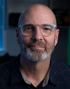 An image of Lee Boot, an adult, light-skinned bald man with a light brown beard who is wearing clear circular glasses and a blue polo shirt.