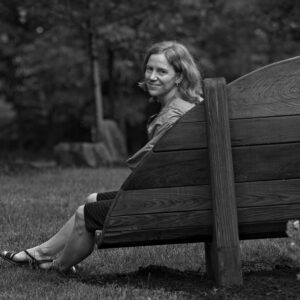A black and white portrait of a smiling adult white woman with light shoulder-length hair. The woman is sitting on a bench in a park with her legs crossed and looking over her left shoulder to smile at the camera. 