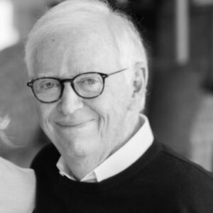 A black and white portrait of a smiling senior white man with white hair who is wearing dark, rounded glasses, and a dark sweater over a white-collared shirt. 