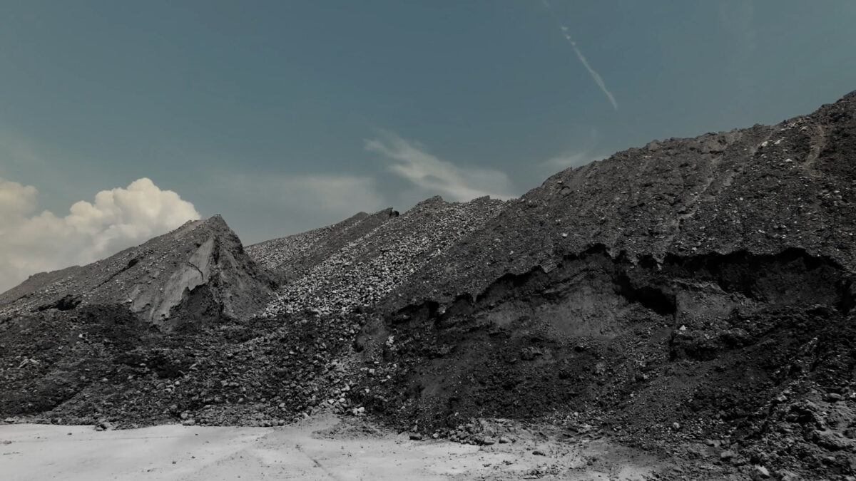Video still from "Conflux: Variation" by Collis Donadio, showing a large grey-colored mound of minerals against a blue sky.