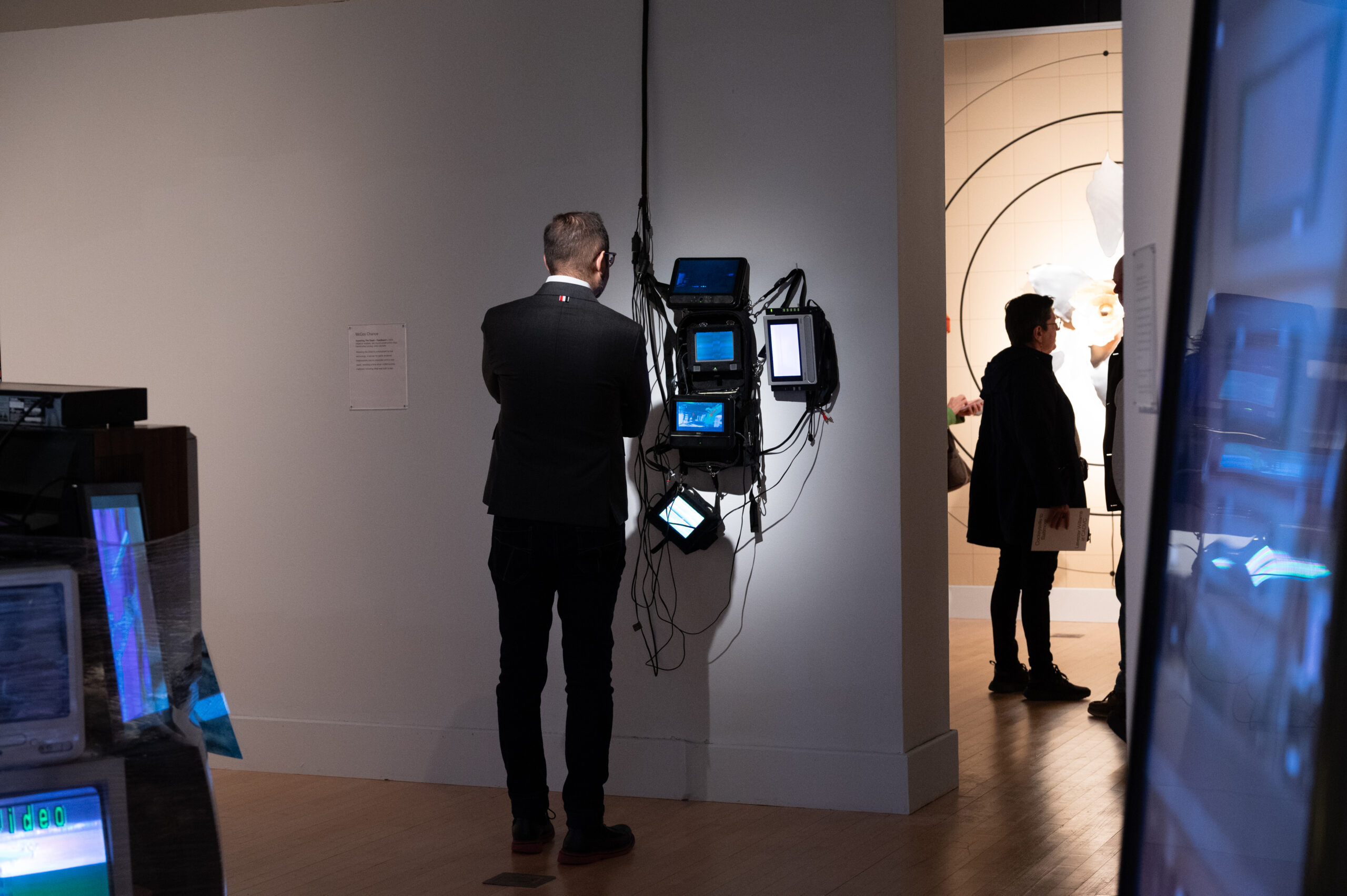 A man in a black suit observes a mixed media installation mounted on the gallery wall. It consists of two monitors and various electronic devices.