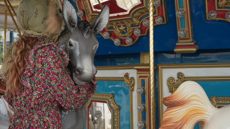 A woman wears a yellow sheer scarf over her face and holds onto a pole on a merry-go-round.