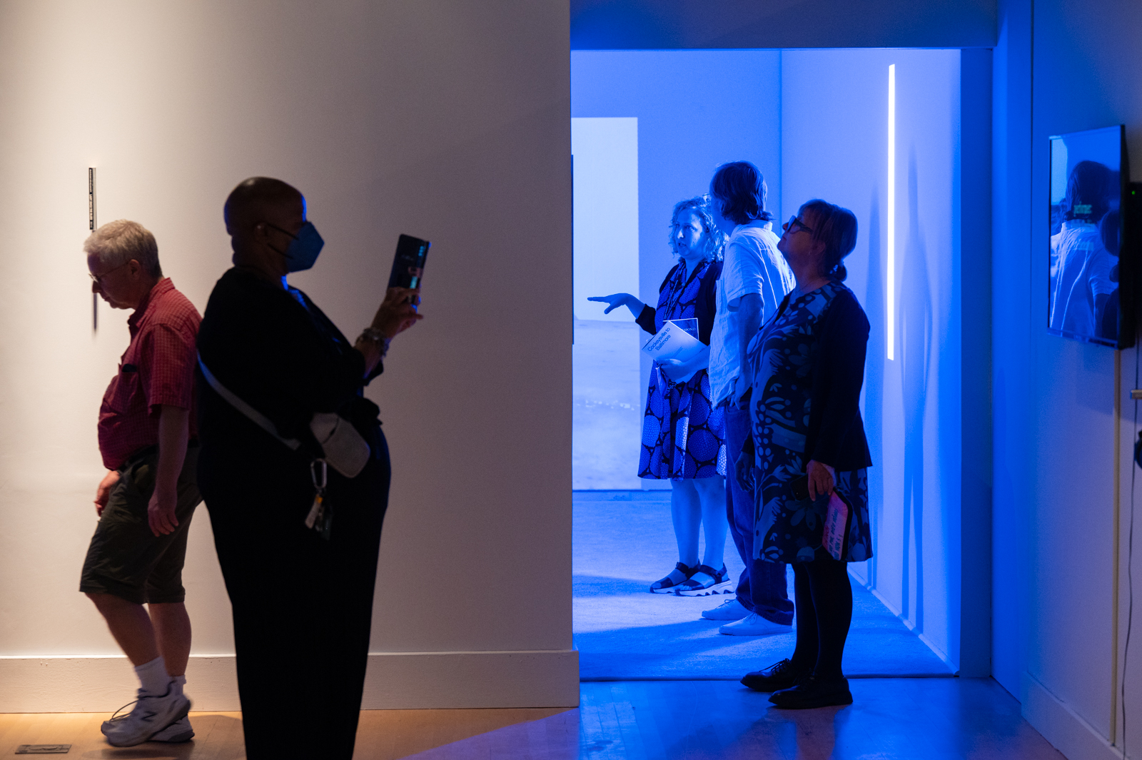 Gallery visitors view works by Levester Williams mounted on the gallery wall. A blue light is emitted from one room of the gallery.