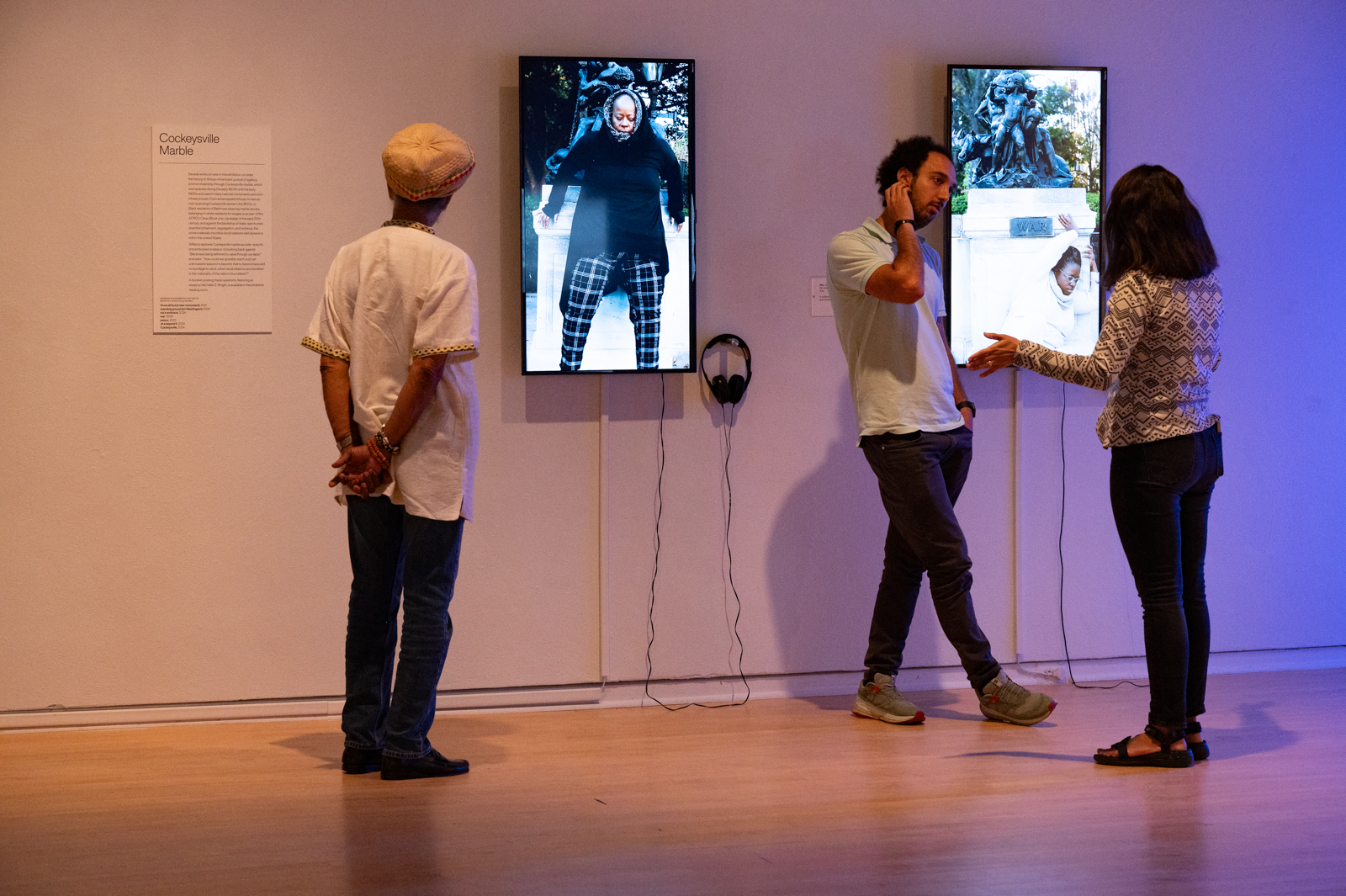 Three individuals in white t-shirts stand near video installations mounted on a gallery wall.