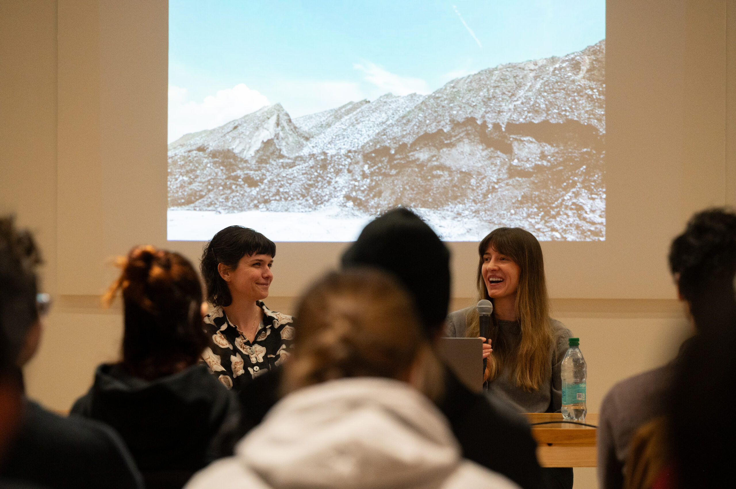 Shannon Collis and Liz Donadio in front of an audience discussing their work, "Conflux: Variation." A still of the work is projected behind them.