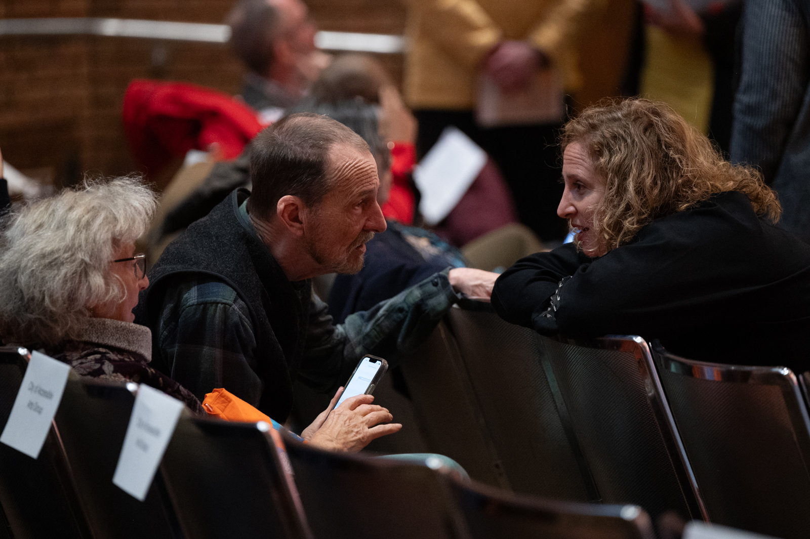 An image of three individuals seated in an auditorium. A woman with blonde curly hair turns around over her chair to speak to two individuals. The man at left leans forward in discussion while the woman at right relaxes in her seat.