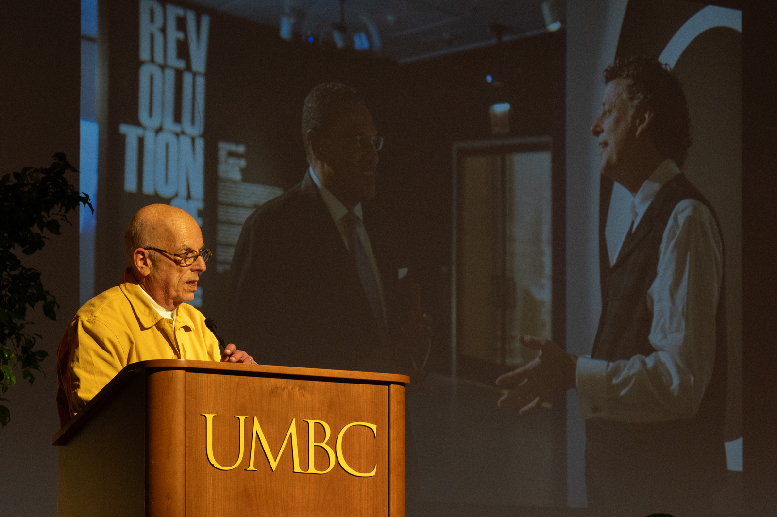 A man with a light skin-tone wearing glasses and a yellow button-down shirt stands at a wooden podium with "UMBC" on the front. Behind him is a projected image of two men speaking in an exhibition.