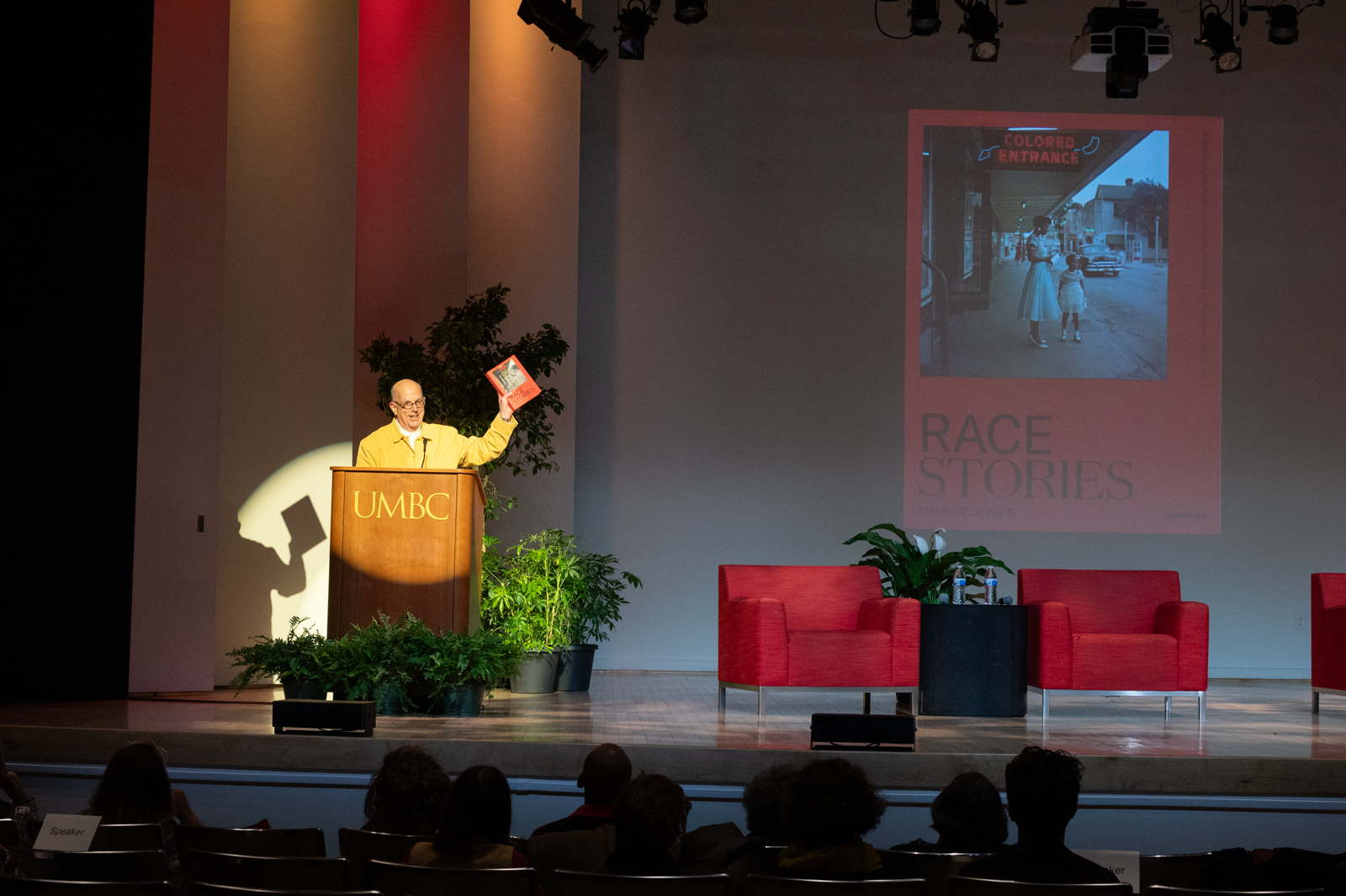 A man with a light skin-tone and wearing a light yellow button-down shirt stands at a wooden podium holding up a red book in his left hand. An image of the book is also projected behind him. A spotlight shines brightly on him and the podium. To his left are two red chairs for a discussion.