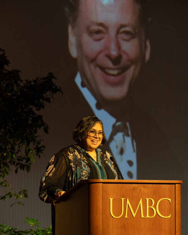 A woman a brown skin-tone with short brown hair stands at a wooden podium with "UMBC" on the front in gold letters. She smiles to the audience.