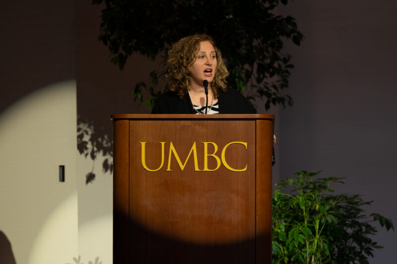 A woman with a light skin-tone and short curly blonde hair stands at a podium with "UMBC" in gold letters. A spotlight shines on her as she speaks.