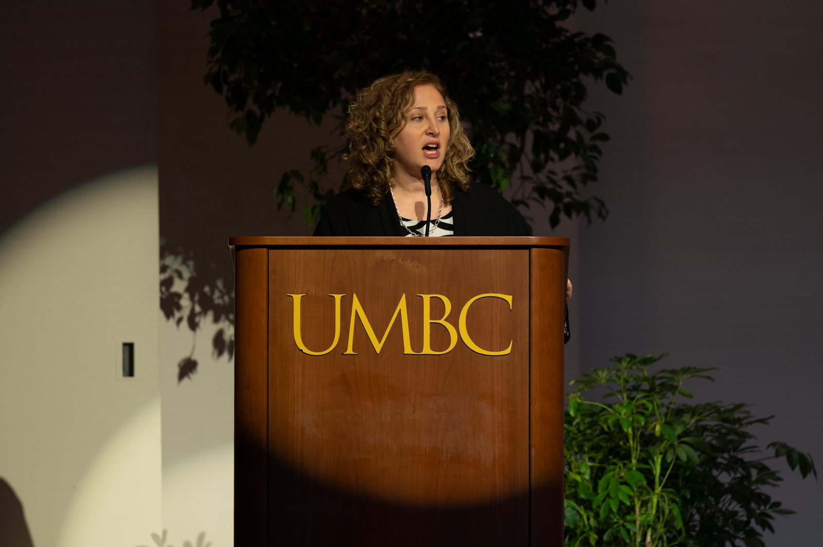 A woman with a light skin-tone and short curly blonde hair stands at a podium with "UMBC" in gold letters. A spotlight shines on her as she speaks.