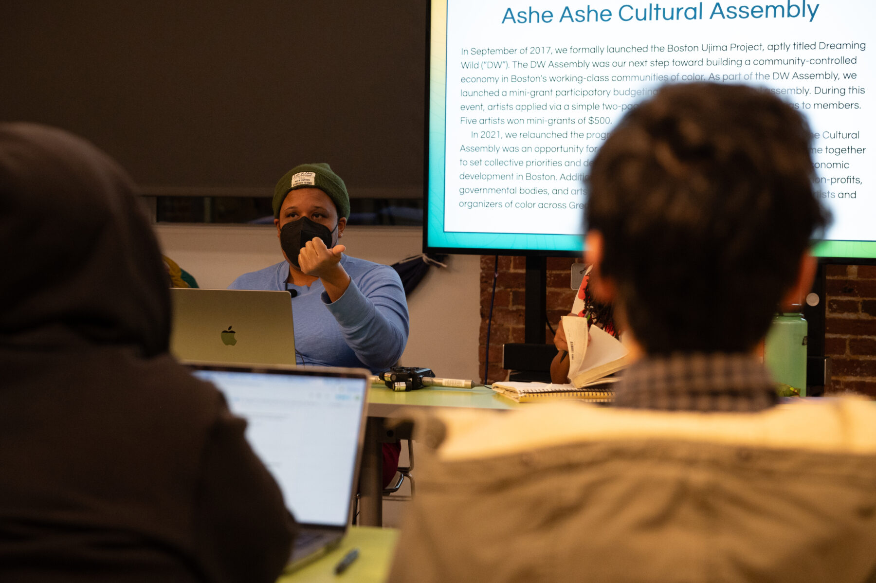 A person with a dark skin-tone. They wear a light blue sweater and a dark green beanie speaks to a group. They are gesticulating with their left hand, pointing her thumb in that direction