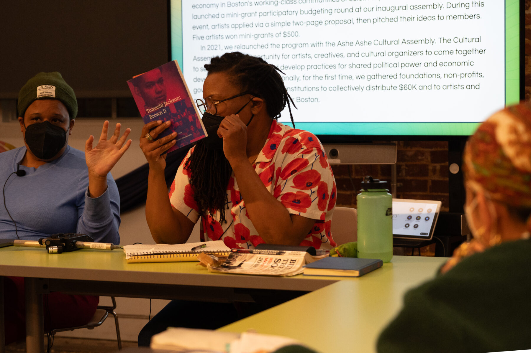 A woman with a dark skin-tone. She wears a black face mask, glasses, and a floral shirt holds up a red duotone book. The title on the cover reads, "Tomashi Jackson: Brown II."