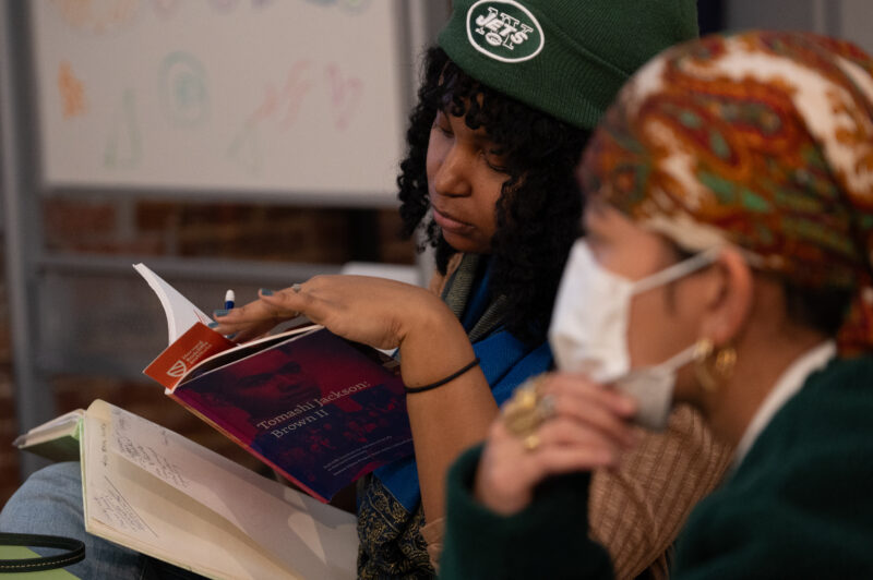 Photo of a person with a dark skin-tone and a New York Jets hat reading from a book. They use their left hand to hold open the book.
