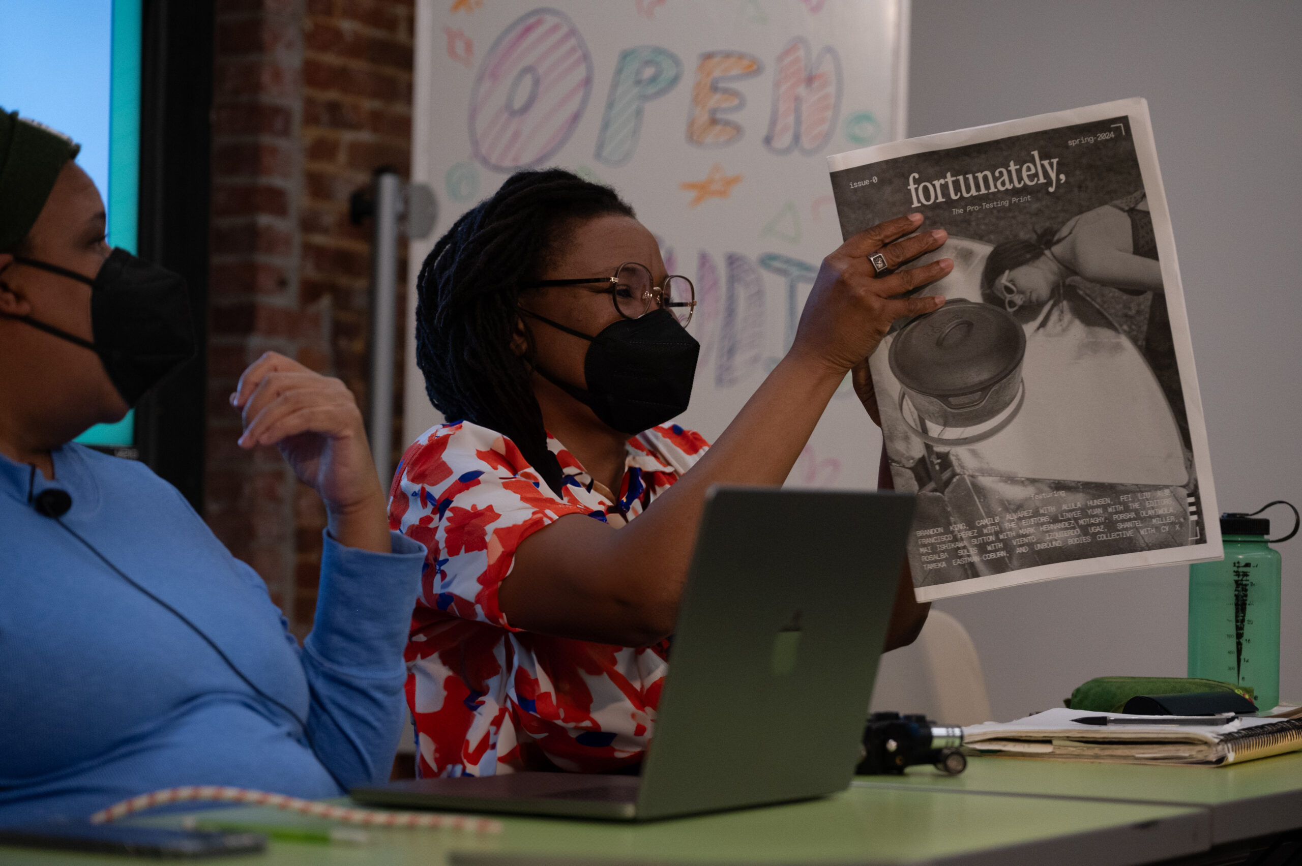A woman with a dark skin-tonewearing a black face mask, glasses, and a floral shirt holds up a newspaper. The front page features a full-page photograph with the title "fortunately."