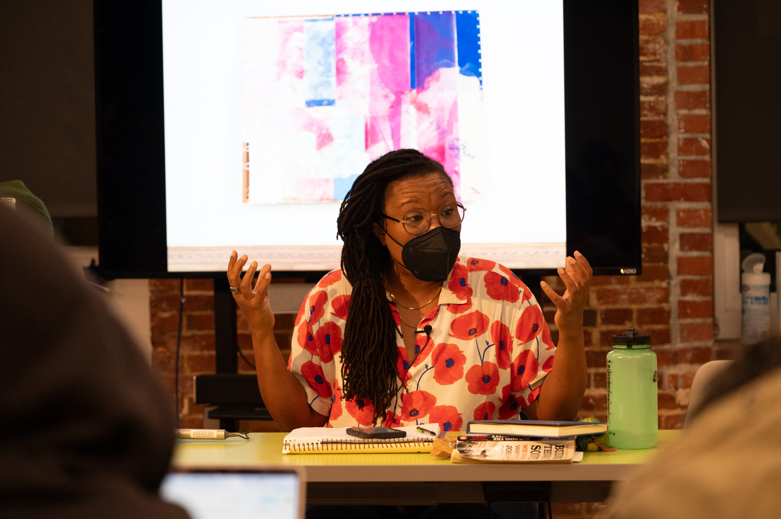 A woman with a dark skin-tone. She wears glasses and a black face mask and is seated at a table in discussion with someone. She is wearing a cream-colored shirt printed with red poppies. She holds up her hands while gesticulating.
