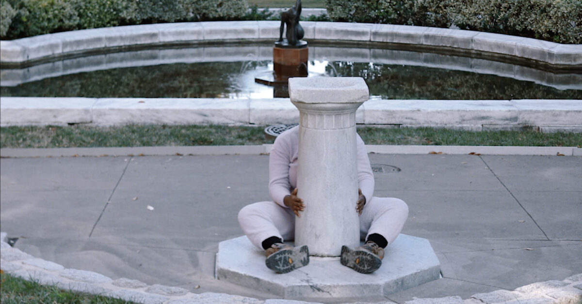 A woman wearing white is seated behind a pillar that rises between her outstretched legs.