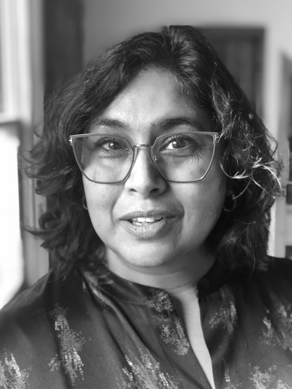 A woman with dark hair and eyes wears glasses and a dark patterned blouse looks forward and smiles.