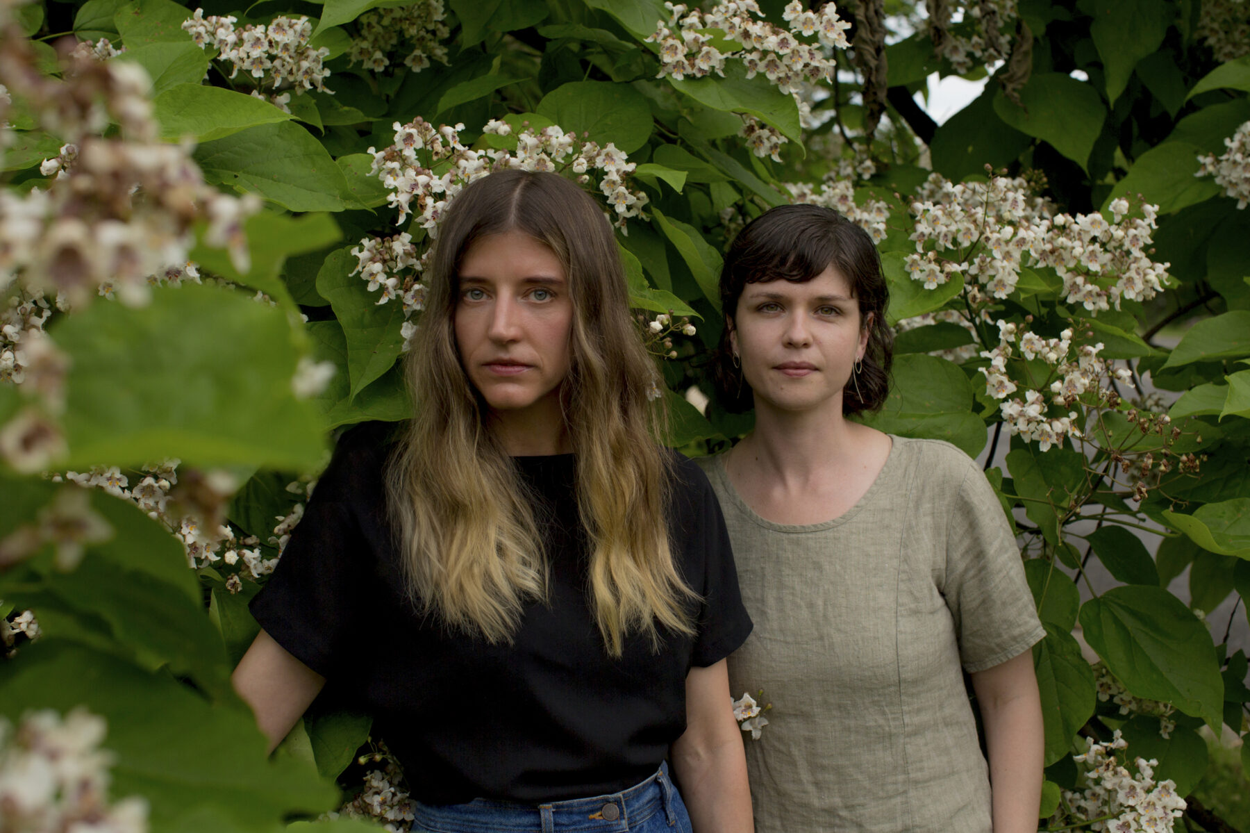Two women stand in front of a flowering bush staring at the camera. The woman at the left has long dirty blonde hair with a black t-shirt while the woman at right has short brown hair and is wearing a grey blouse.