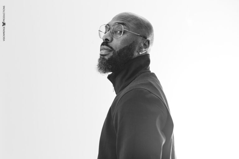 Black-and-white headshot of a man. He has a shaved head and a full beard, wearing round glasses and a black turtleneck. He is looking over his left shoulder at the camera.