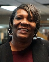Photo of a woman smiling at the camera. She has brown hair in a pixie cut with gold hoop earrings. She wears a dark blazer and a maroon red shirt.