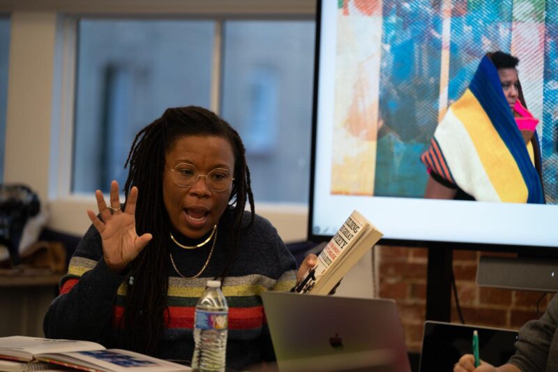 A Black woman reads emphatically from a book open in her left hand. She gesticulates with her right hand.
