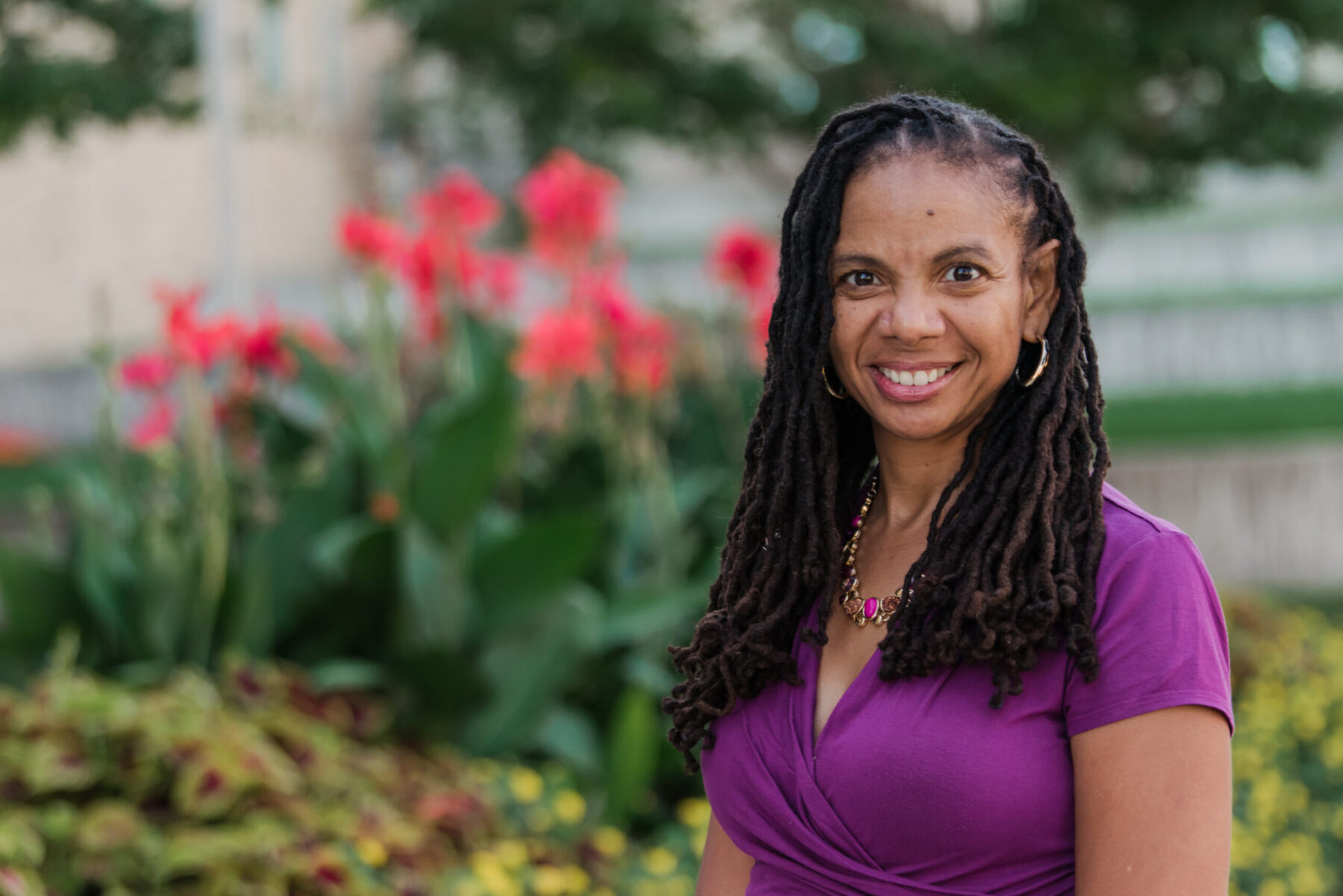 Kimberly Moffitt, a woman with light brown skin tone and long dark hair wears a purple blouse, hoop earrings, and smiles while looking forward.