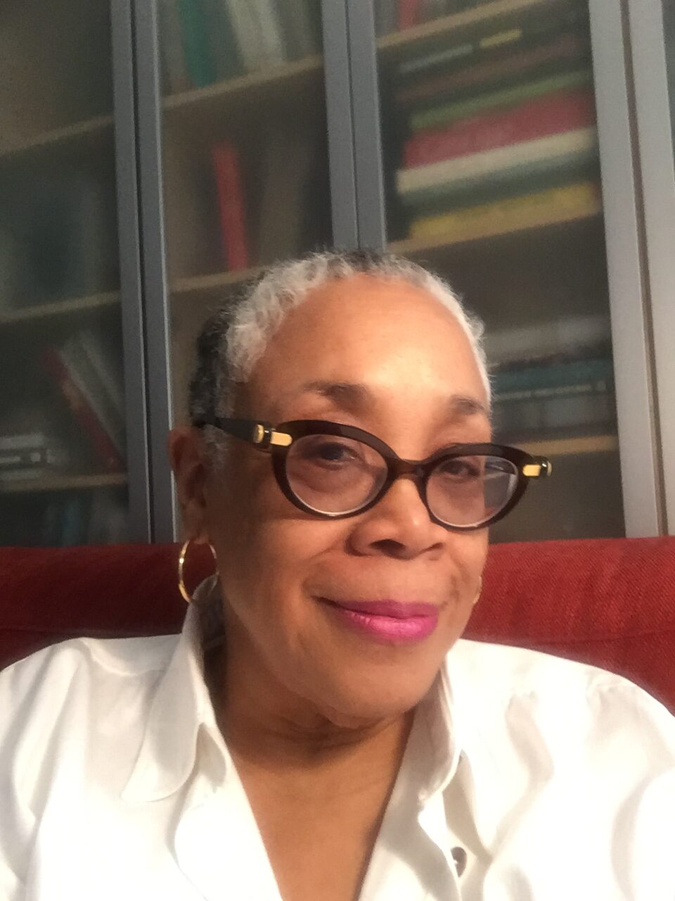 Lowery Stokes Sims, a woman with light brown skin tone wears glasses, a white shirt, golden earrings, and lipstick and smiles, looking forward at an angle.