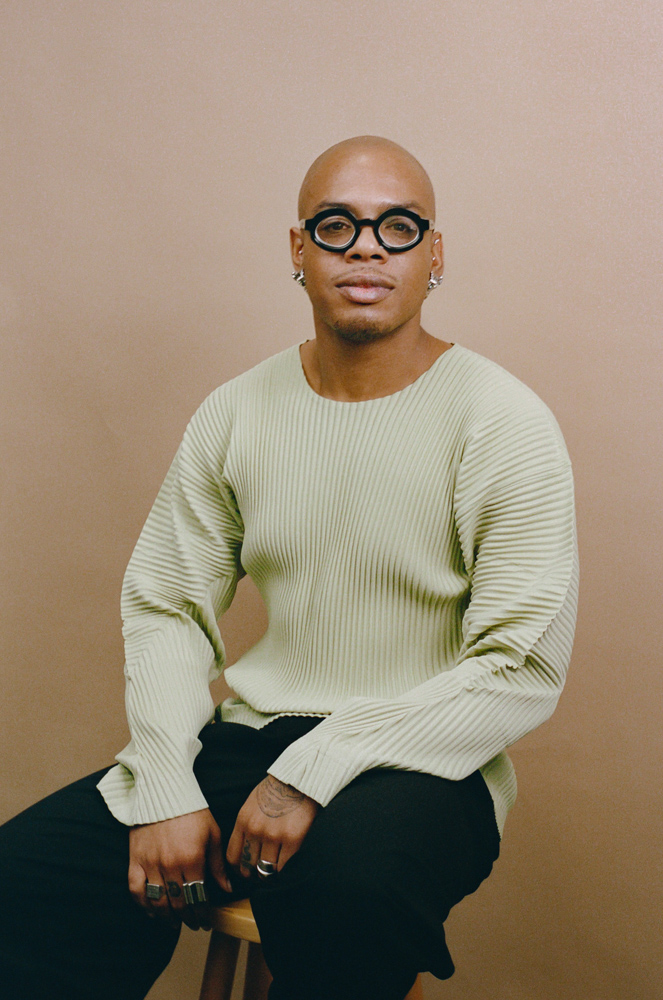 Meleke Glee, a man with a bald head, a thin moustache, light brown skin tone, and round, black-rimmed glasses wears a light sweater and sits on a stool and looks forward.
