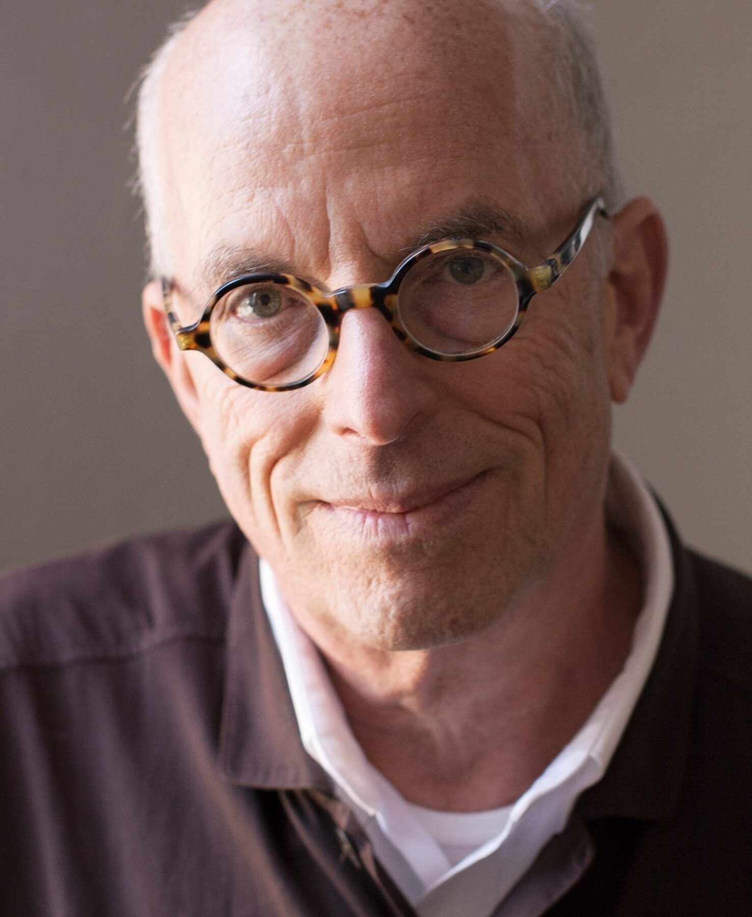 Marvin Heiferman, a man with a light complexion smiles and wears round-framed glasses and dark collared shirt layered over a white collared shirt and a white t-shirt.