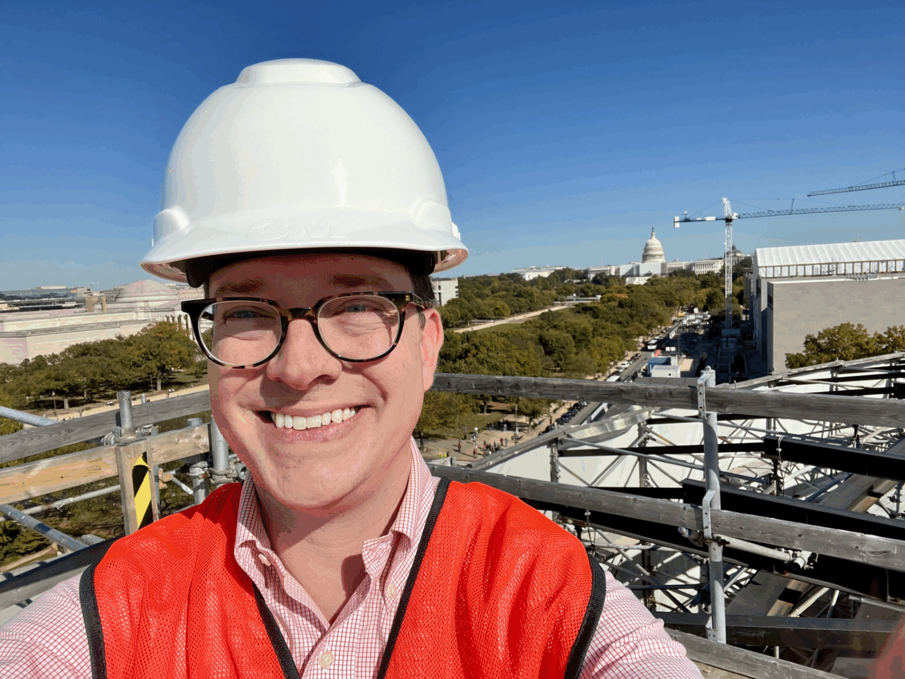 Photo of a man with glasses smiling at the camera. He is wearing a white hard hat and a high vis vest.