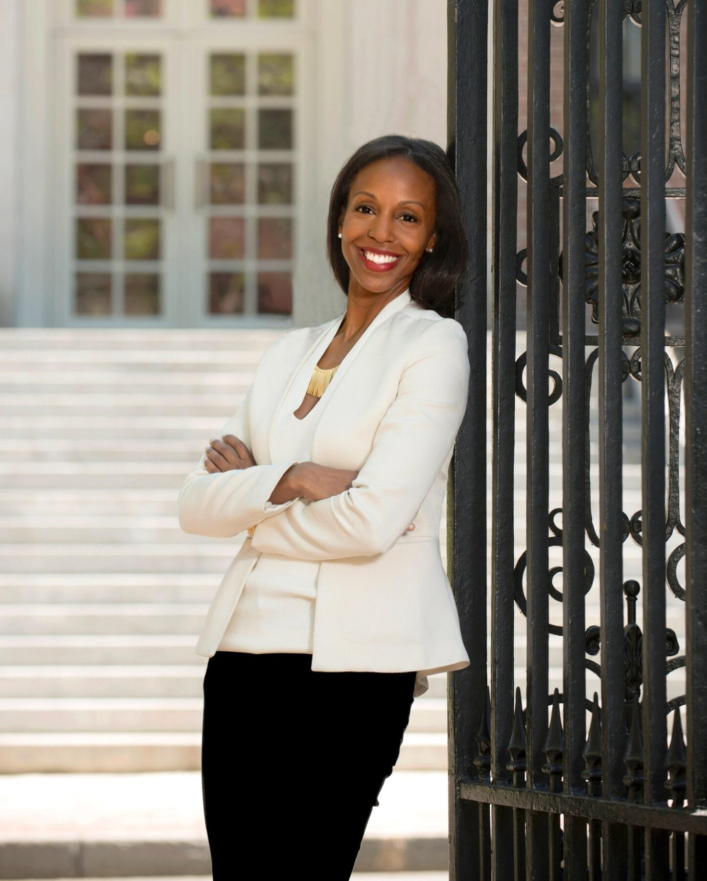 Sarah Lewis, a woman with medium brown skin tone, wears a white suit jacket and smiles with arms crossed as she leans against a wrought iron gate and looks forward.