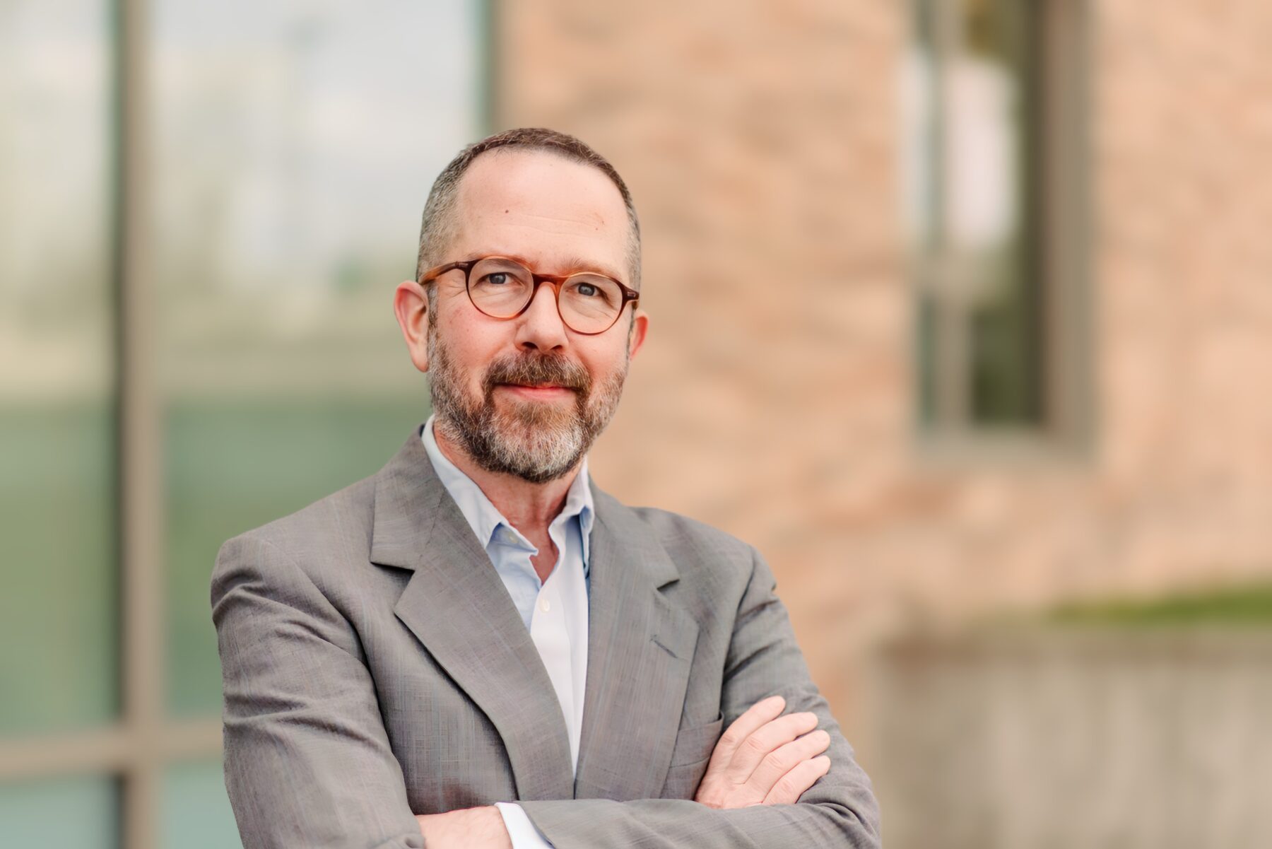 Timothy Nohe, a man with a light skin tone and a beard wears glasses and a grey suit jacket and smiles with his arms crossed as he looks forward.