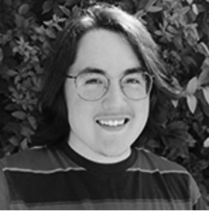A black-and-white photo of a man with medium length dark hair smiling at the camera. He wears round wireframe glasses and a striped shirt.