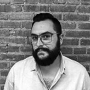 A black-and-white photo of a dark-haired bearded man looking at the camera. He wears a white button-down shirt, unbuttoned at the collar, with dark-rimmed glasses. He stands against a brick wall.