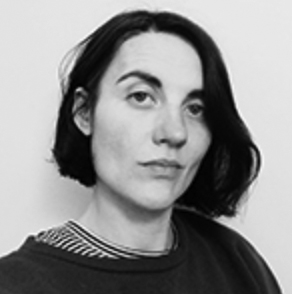 A black-and-white photo of a woman looking at the camera. Her dark-haired bob covers one eye as she tilts her head to the left. She wears a dark shirt over another a striped shirt.
