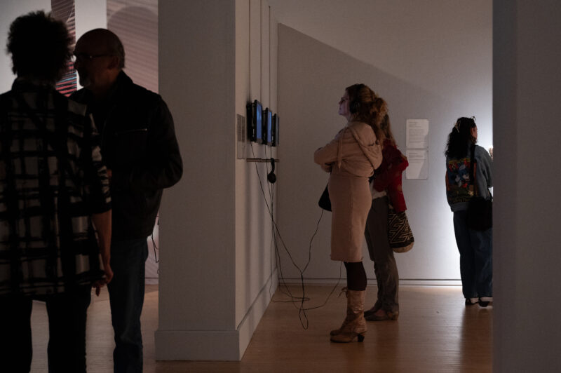 Two women observe a mixed media installation mounted on the wall of the gallery.
