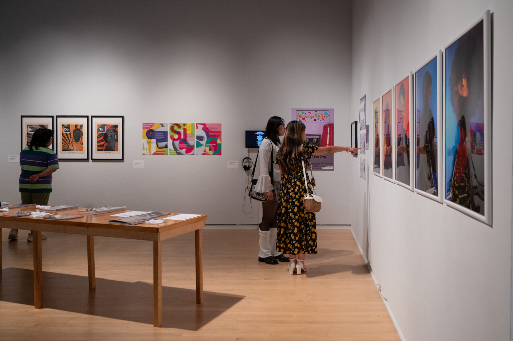 Two women stand in a gallery space in which framed works hang on the walls. One woman with brown hair and a floral printed dress points at something in one of these works, while her companion in a white outfit observes.