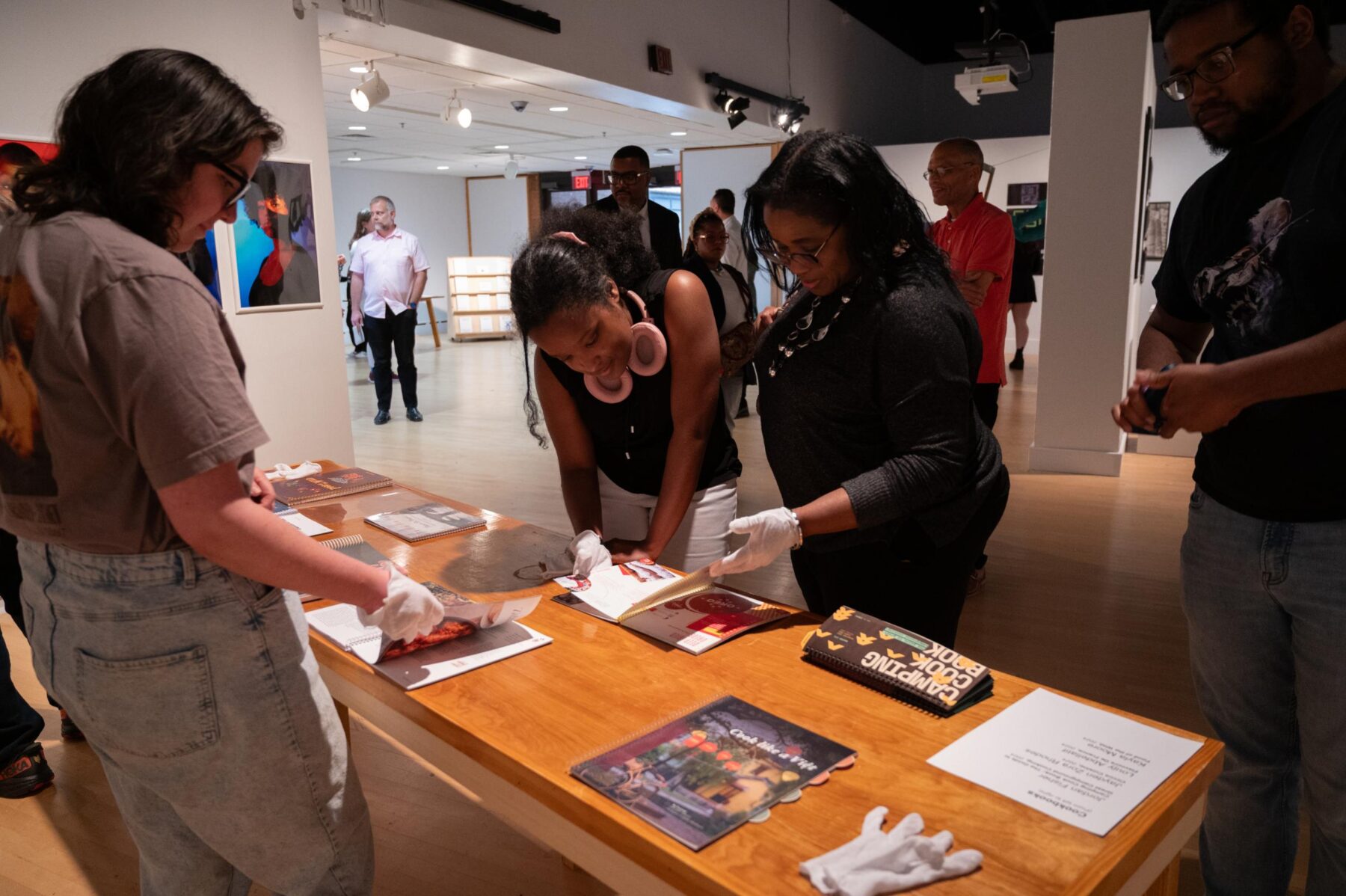 A group of gallery visitors flip through publications on a wood table. All are wearing white gloves to handle the printed matter.