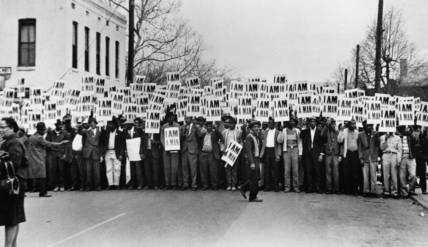 A black-and-white photograph featuring a large group of Black men standing on a street carrying picket signs. The signs read "I Am A Man" in capital letters. In the background is a white two story building and electrical poles, which line the street they stand upon.