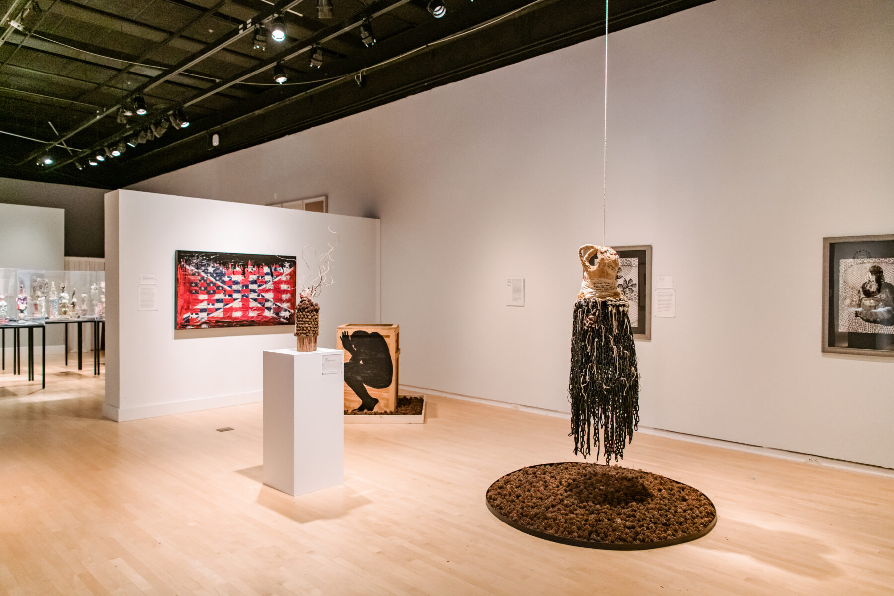 Installation view of Oletha DeVane: Spectrum of Light and Spirit. A mixed media art work resembling a dress hangs from the ceiling above a circular work on the wooden floor.