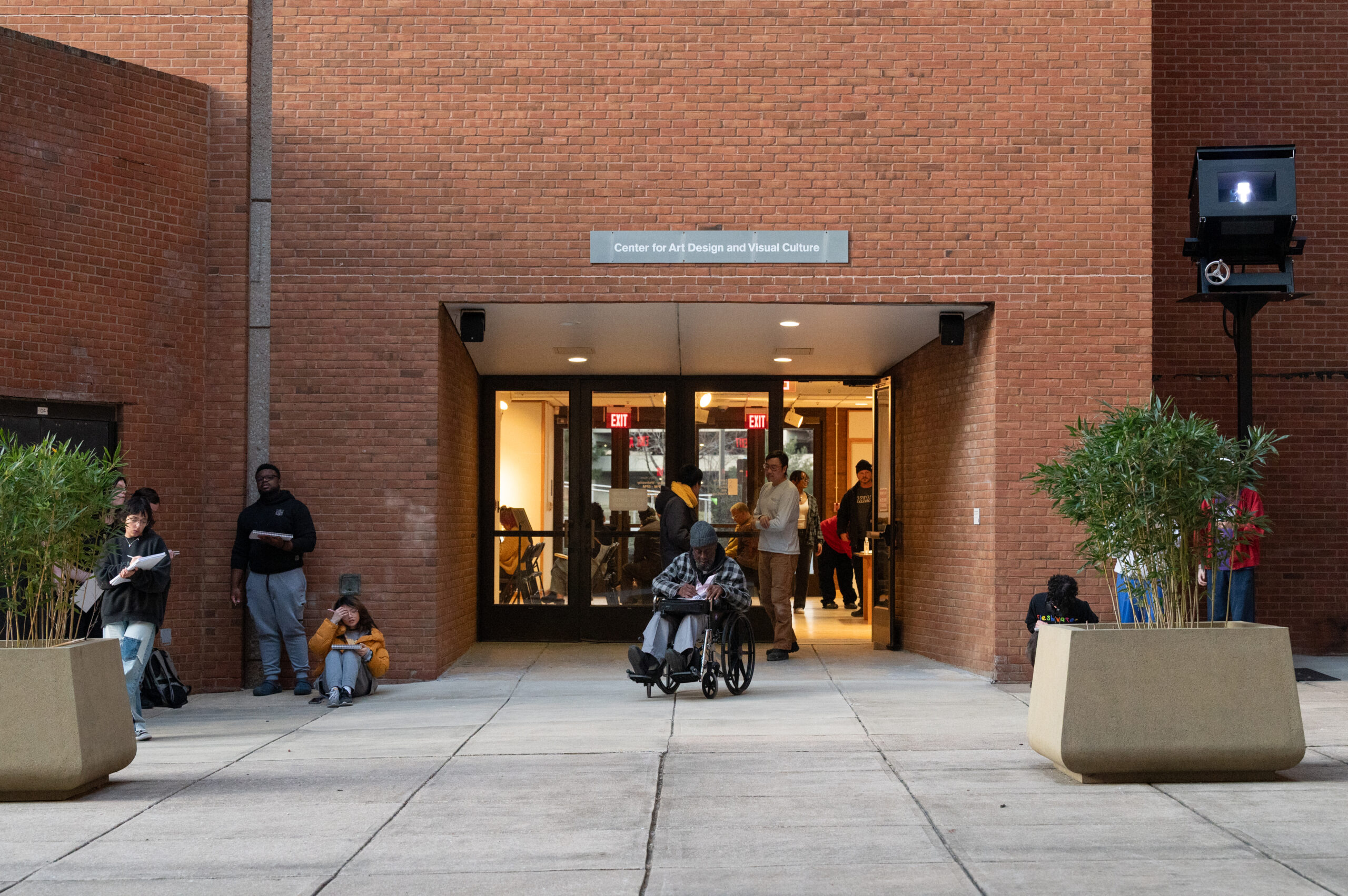 Exterior photo of a red brick building and a double door entrance. A man in a wheelchair is seated at center with a plaid blanket shawl over his shoulders.