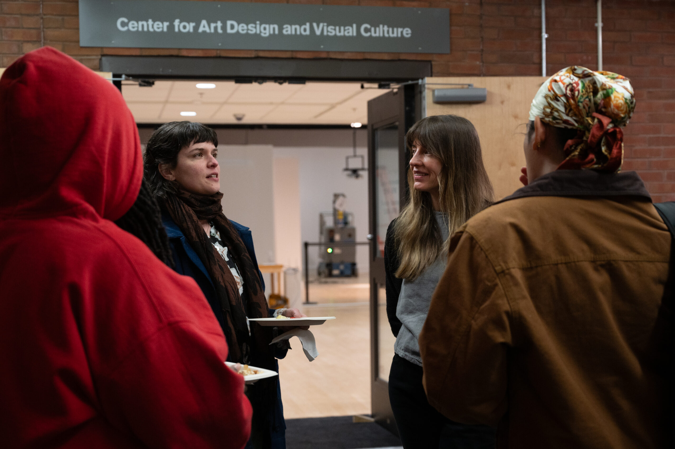Four individuals stand together outside in discussion. At center are two women with light skin-tones speaking to their companions.