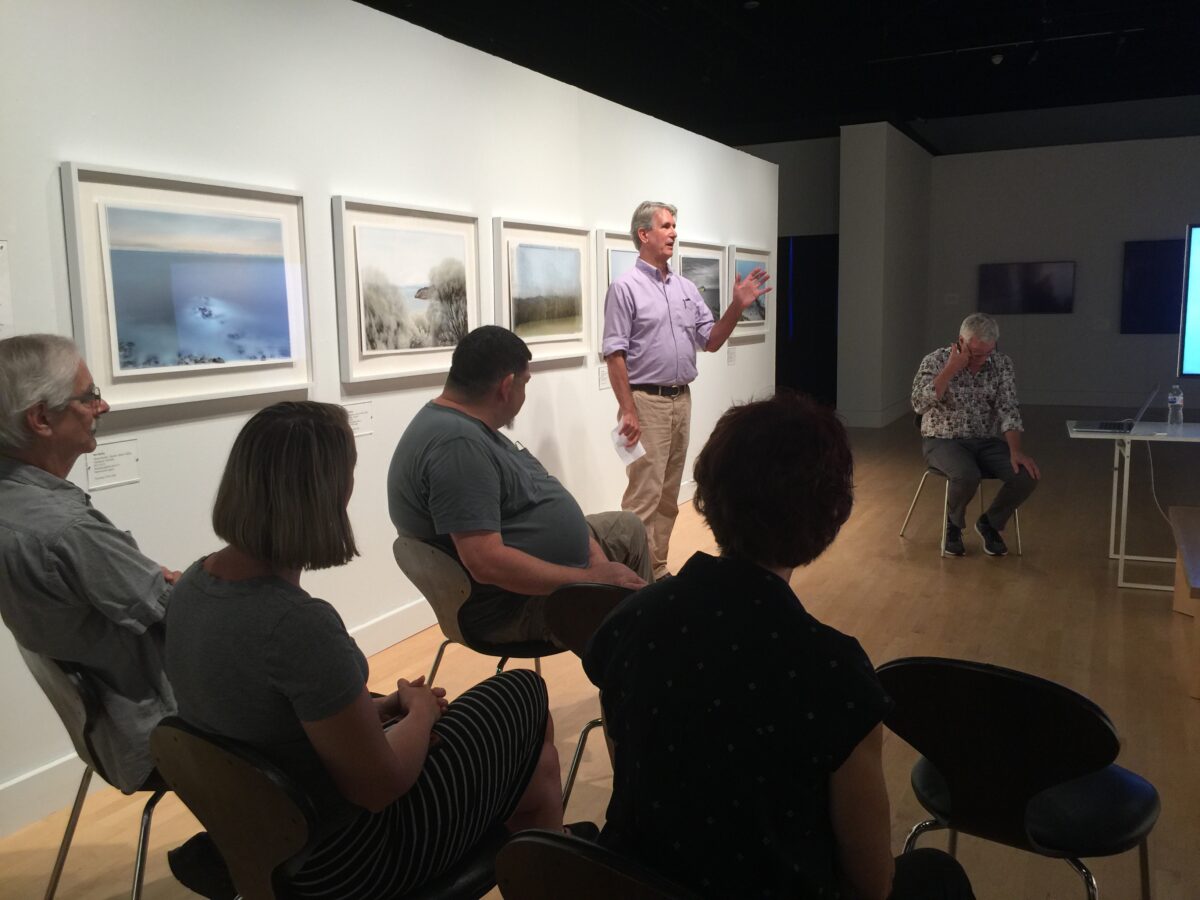 A light-skinned man in a lavender button down stands in front of framed works on a white gallery wall. He is speaking to a larger audience, who is seated in front of him in silhouette.