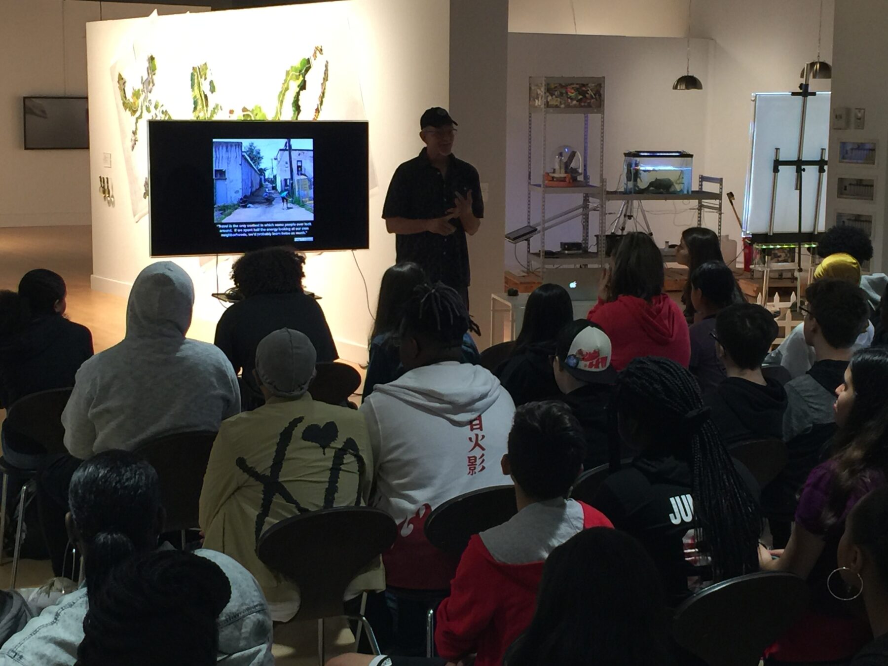 A man in silhouette stands in front of a large audience seated in a gallery. Behind him is a monitor screen.