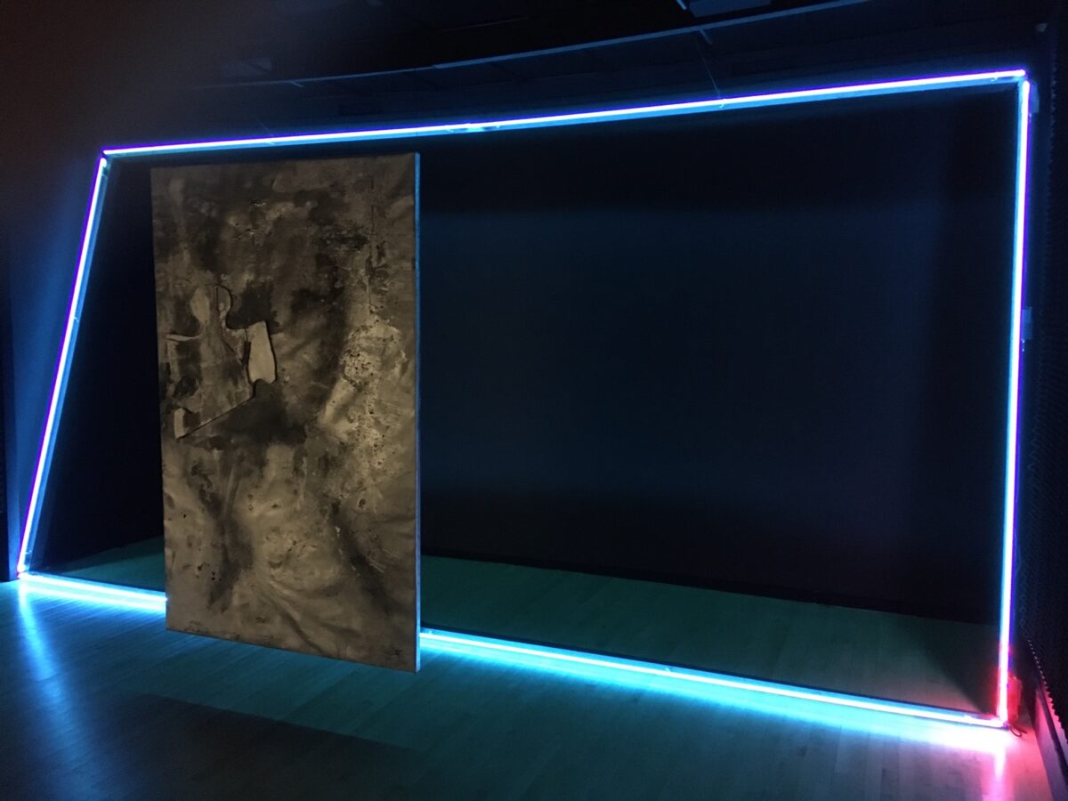 Installation view of a silver metallic abstract sculpture to the left side of a neon frame. The neon shines blue, red, and green in an otherwise dark room.